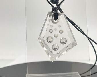 May include: Clear, geometric pendant necklace with a black cord. The pendant is a diamond shape with embedded clear bubbles of varying sizes. The necklace is displayed against a white background.