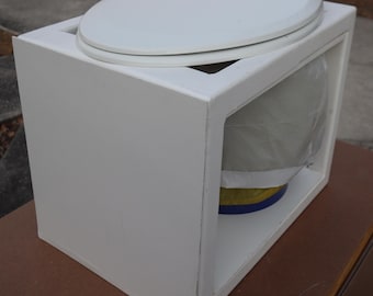 May include: A white toilet bowl with a white lid, enclosed in a white box with a clear front panel. The toilet bowl is partially visible through the panel.