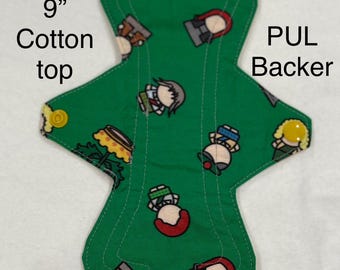 Può includere: Un assorbente igienico riutilizzabile verde con una stampa di cartoni animati. L'assorbente misura 22,86 cm di lunghezza e ha un top in cotone e un retro in PUL. Il testo sull'assorbente dice "Assorbimento regolare".