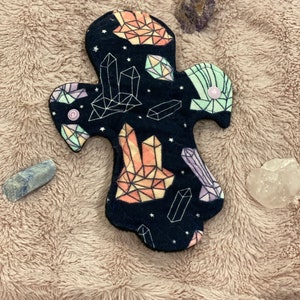May include: A reusable cloth menstrual pad featuring a dark blue fabric with a geometric crystal pattern in peach, green, and purple. The pad has a button fastening and is surrounded by crystals on a textured, light brown surface.