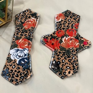 May include: Two reusable cloth menstrual pads with a leopard print and floral design. The pads are brown, red, white, and black.