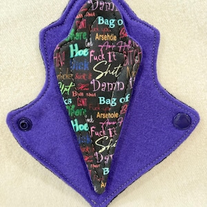 Puede incluir: Una compresa menstrual de tela reutilizable con un exterior morado y un interior negro. El interior negro está cubierto de texto colorido, incluyendo las palabras "Damn", "Shit" y "Fuck It". La compresa tiene cierres de presión.
