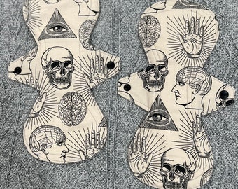 May include: Two white cloth menstrual pads with a black and white print of skulls, eyes, brains, and hands. The pads are on a gray knitted surface.