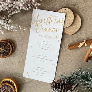 Op de afbeelding: Een wit kerstdiner-menu met gouden letters en steraccenten. Het menu bevat voorgerechten, hoofdgerecht en desserts. Decoratieve elementen zijn houten schijfjes, kaneelstokjes en gedroogde sinaasappelschijfjes.