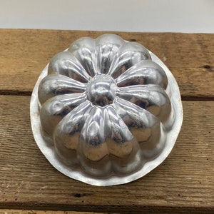 May include: A silver metal mold for making a flower-shaped cake or gelatin dessert. The mold has a fluted design and a round center.