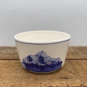 May include: A white ceramic planter with a blue and white windmill scene. The planter is round and has a wide rim.