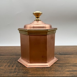 Peut inclure: Un récipient octogonal de couleur cuivre avec un couvercle et un bouton doré. Le récipient a une garniture de couleur laiton autour du couvercle. Le récipient est posé sur une surface en bois.