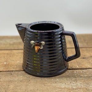 May include: A black ceramic jug with a bee-shaped decoration. The jug features a ribbed design and a handle. The bee is brown, yellow, and white. The jug is on a wooden surface.