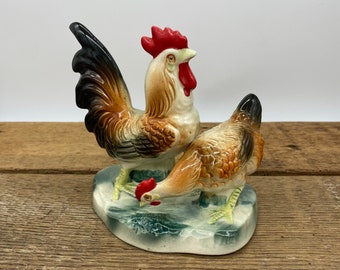 May include: A ceramic rooster and hen figurine set. The rooster is brown, black, and red with a red comb. The hen is brown and white. The figurines are standing on a green and white base.