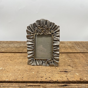 May include: A small, ornate silver-colored picture frame with a rectangular opening. The frame has a ruffled edge and is decorated with small, gold-colored floral accents at the top and bottom.