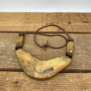 May include: A wooden necklace with a boomerang-shaped pendant and brown leather cord. The pendant has a natural wood grain pattern with brown and yellow tones.