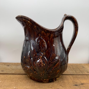 May include: A brown ceramic pitcher with a floral design. The pitcher has a wide mouth and a handle.