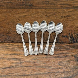 May include: A set of six silver-toned metal spoons arranged in a row. Each spoon features a smooth, oval-shaped bowl and a slender handle with a subtle decorative pattern at the base. The spoons have a polished, reflective finish throughout.