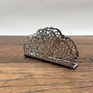 May include: A silver-colored metal napkin holder featuring an intricate, openwork filigree design with a scalloped top edge. The structure is composed of thin, curved metal strips forming a decorative pattern that allows for holding napkins upright.