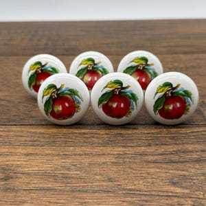 May include: Six white ceramic cabinet knobs with a red apple design. Each knob features a detailed illustration of a ripe red apple with green leaves and a blue background. The knobs are arranged on a wooden surface.