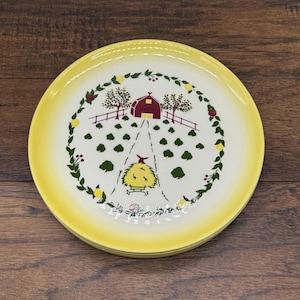 Peut inclure: Une assiette ronde en céramique avec un bord jaune vif et un centre blanc. Le motif présente une illustration centrale d'une grange rouge, d'arbres verts et d'une meule de foin jaune sur un chemin, entourés d'une couronne de feuilles vertes.