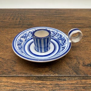 May include: A ceramic candle holder featuring a white base with blue hand-painted stripes and abstract markings. The circular tray has a central cylindrical socket for a candle and a small, rounded handle on the side, all finished in a glossy glaze.