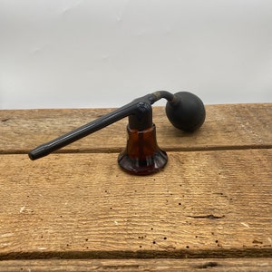May include: A vintage glass atomizer with a black rubber bulb and a long black nozzle. The atomizer is sitting on a wooden surface.