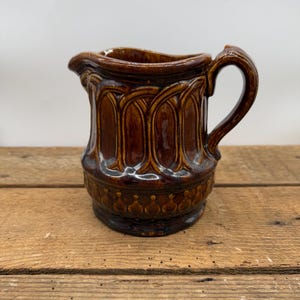 May include: A brown ceramic pitcher with a decorative design. The pitcher has a rounded body and a handle.