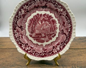 May include: A decorative plate with a scalloped edge, featuring a burgundy and white design. The center showcases a scenic landscape with trees and buildings. The rim is adorned with a burgundy leaf pattern. The plate is displayed on a gold-colored stand.