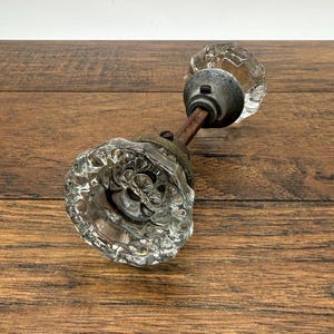 May include: A vintage glass door knob with a clear, faceted design. The knob is attached to a metal rod and mounting hardware with a tarnished finish. The door knob is a classic architectural element.