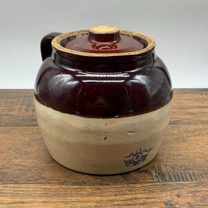 May include: A two-toned stoneware crock with a dark brown glazed upper section and a natural beige lower section. It features a matching dark brown lid with a textured rim and a blue stamped logo on the front containing the number 2.