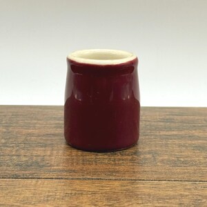 May include: A small, cylindrical, burgundy-colored ceramic jar with a cream-colored interior rim. The jar is set on a wooden surface, and the background is plain white. The jar has a glossy finish.