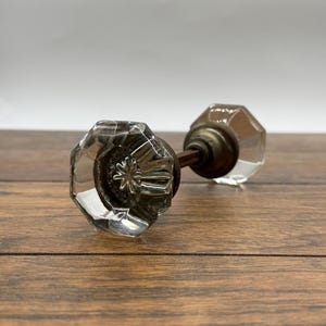 May include: Clear glass octagonal door knobs with a bronze-colored metal base and stem. The knobs are resting on a wooden surface. The glass has a faceted design, reflecting light. The hardware is likely vintage or antique.