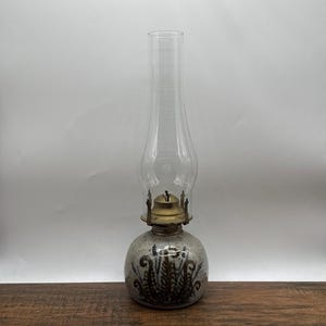 May include: A vintage oil lamp with a clear glass chimney and a ceramic base. The base is decorated with a brown floral pattern. The lamp has a brass burner and a clear glass chimney. The lamp is sitting on a wooden surface.