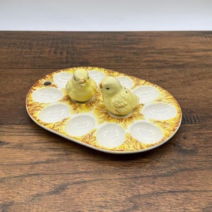 May include: An oval ceramic egg holder with 12 egg slots, decorated with yellow and brown accents. Two yellow ceramic chick figurines sit on top. A charming Easter or spring-themed kitchenware item.