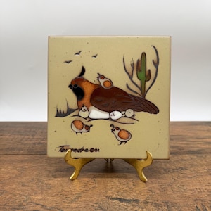 May include: A square ceramic tile with a cream-coloured background featuring a brown quail with chicks and a green cactus. The illustration includes small black birds in the sky and the signature "Tessedre ©82" in the bottom left corner.