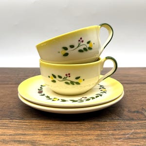 May include: Two cream-colored ceramic teacups and two matching saucers with a yellow rim. The cups and saucers feature a hand-painted design of green leaves, small yellow berries, and tiny red dots. One cup is stacked on top of the other cup.