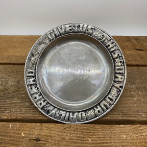 May include: A silver metal dish with a raised rim and the words "Give Us This Day Our Daily Bread" inscribed around the edge.