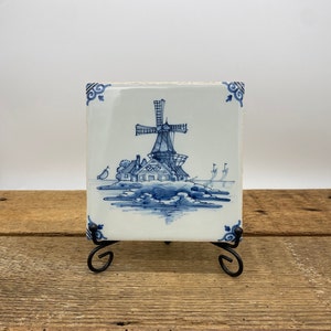 May include: A blue and white Delft tile depicting a windmill and a house with a blue and white painted design. The tile is on a black metal stand.