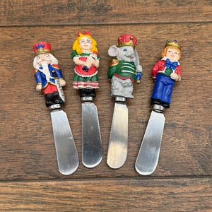May include: Four decorative butter knives with character-themed handles. The handles feature a nutcracker, a girl in a green dress, a mouse in a green jacket, and a boy in a red jacket. The blades are silver-colored.