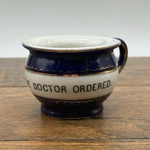May include: A small ceramic chamber pot mug featuring a deep blue and white color scheme with gold trim accents. The white center band displays the black text "THE DOCTOR ORDERED." The vessel has a rounded body, a flared rim, and a single side handle.
