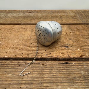 May include: A silver metal tea infuser with a chain and hook. The infuser is shaped like a bell and has a perforated design.