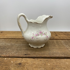 May include: A white ceramic creamer with a pink floral design. The creamer has a scalloped rim and a handle.