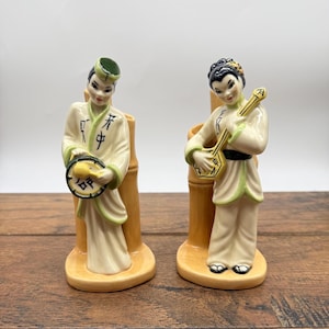 May include: A pair of vintage ceramic figurines depicting Asian musicians. Each figure is dressed in cream-colored robes with green trim and holds a musical instrument. The figurines are mounted on a bamboo-style base and are in excellent condition.