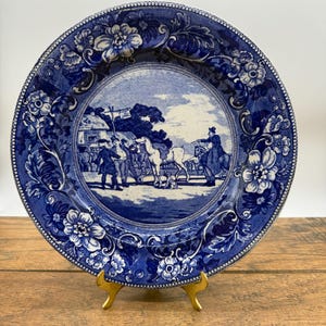 May include: Blue and white decorative plate with a pastoral scene in the center, surrounded by floral and scrollwork patterns. The plate is displayed on a gold-colored stand, showcasing its intricate design and vintage aesthetic.