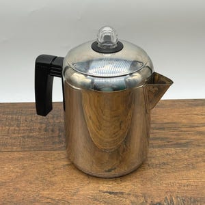 May include: A polished stainless steel percolator coffee pot featuring a cylindrical body, a black heat-resistant handle, and a domed lid topped with a clear glass knob. The coffee pot includes a triangular spout and a smooth, reflective metallic finish.