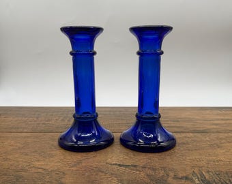 May include: Two cobalt blue glass candle holders. Each holder has a flared base, a cylindrical stem with a decorative ring, and a flared top. The candle holders are displayed on a wooden surface.