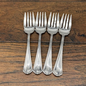 May include: A set of four silver-toned metal forks arranged side by side. Each fork features a four-tine head and a slender handle that widens toward the base, displaying a subtle engraved monogram detail near the bottom of the handle.