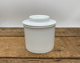 Könnte beinhalten: Eine weiße Keramik-Butterdose mit Deckel. Die Dose ist rund und hat eine glatte, glänzende Oberfläche.
