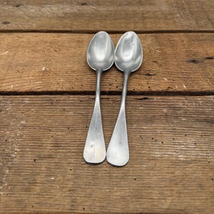 May include: Two vintage silver spoons with elongated handles and rounded bowls, arranged side-by-side. The spoons rest on a rustic, weathered wooden surface, showcasing their aged appearance and classic design. The spoons are made of metal.