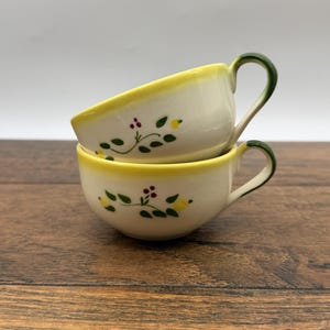 Peut inclure: Deux tasses en céramique empilées avec une base de couleur crème, ornées d'un motif peint à la main de petits citrons jaunes, de feuilles vertes et de minuscules baies rouges. Chaque tasse a un bord jaune vif et une anse verte incurvée.