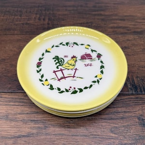 Peut inclure: Une pile d'assiettes rondes en céramique avec un centre blanc et un bord jaune. Le centre présente une couronne de feuilles vertes entourant un coq jaune derrière une clôture rouge, avec une petite grange rouge et des feuillages verts.
