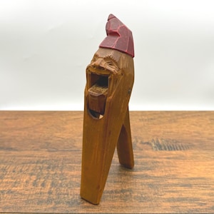 May include: A wooden nutcracker carved in the shape of a man's face with a red hat. The nutcracker has a hinged jaw that opens and closes to crack nuts.