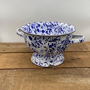 May include: A white enamel colander with blue speckles and two handles. The colander has a round base and is made of metal.
