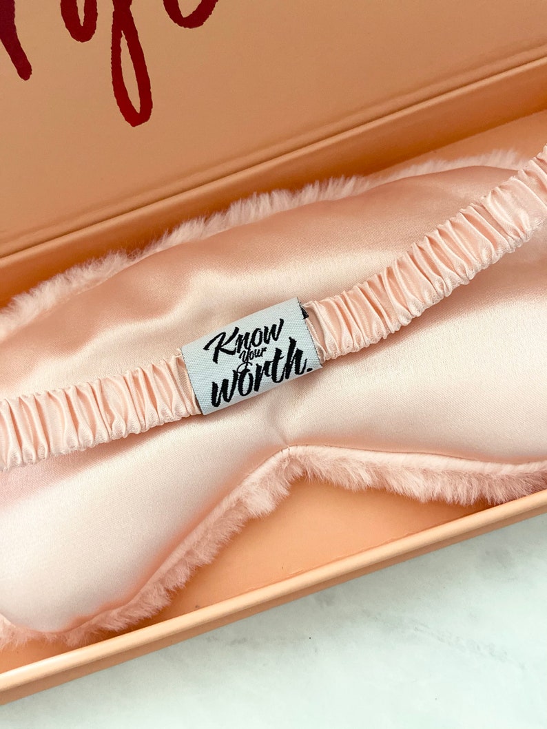 Może przedstawiać: R&oacute;żowa satynowa maska do spania z puszystym białym wykończeniem. Maska ma białą etykietę z napisem "Know your worth".
